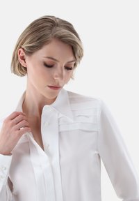 Camicia bianca realizzata in tessuto morbido con colletto strutturato, caratterizzata da pieghe orizzontali sul petto e dettagli con bottoni sui polsini.