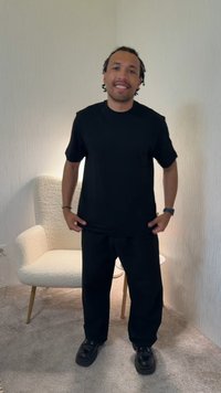 Άνδρας με πλεξούδες που χαμογελάει ενώ στέκεται, φοράει μαύρο t-shirt και παντελόνι, με μια λευκή πολυθρόνα και ένα μικρό τραπέζι στο φόντο.