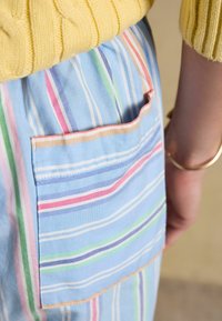 Gros plan sur une poche de jupe rayée bleue, rose, verte et blanche avec une manche de pull côtelé jaune et un bracelet en or au bras.