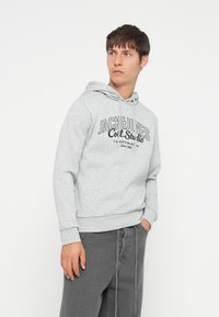 Sudadera gris de tela suave con un estampado de logo en el frontal. Tiene capucha con cordones y puños y dobladillo acanalados, combinada con pantalones gris oscuro.