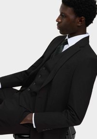 Młody mężczyzna w czarnym garniturze trzyczęściowym i ciemnym krawacie, siedzący w profilu na tle jednolitego jasnego tła, z rękami w kieszeniach.