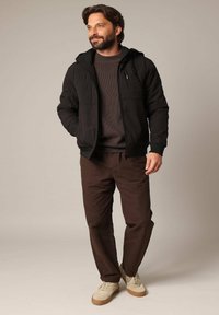 Zwarte ritsjack met capuchon, grijze gebreide trui, bruine broek en beige sneakers. Model staat casual met de handen in de zakken.