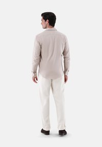 Camicia button-up beige chiaro con yoke posteriore e maniche lunghe, abbinata a pantaloni bianchi e scarpe marroni scure. Tessuto dalla consistenza liscia.