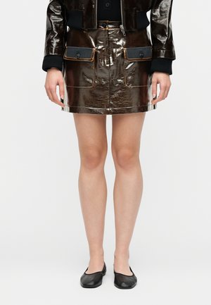 Skinnende mørkebrun lædermininederdel med frontlommer med klap, båret med sorte flade sko og en sort jakke med ribstrikkede manchetter.