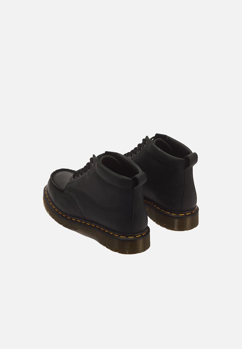 Paar schwarze Leder-Stiefeletten mit gelber Naht und durchscheinend braunen dicken Gummisohlen, von hinten auf weißem Hintergrund gesehen.