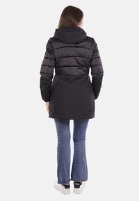 Giubbotto imbottito nero con cappuccio, con una parte superiore trapuntata e una parte inferiore liscia, abbinato a jeans a zampa d'elefante blu e sneakers bianche.