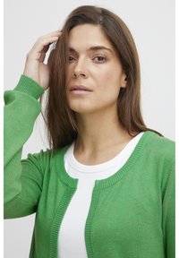 Cardigan en tricot vert avec un col rond, des poignets côtelés et une ouverture à l'avant. Porté sur un t-shirt blanc à col rond. Texture lisse.