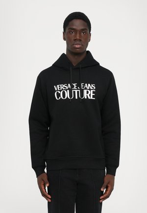 Black hoodie with white "VERSACE JEANS COUTURE" text worn by a man standing against a plain white background, hands relaxed by sides.