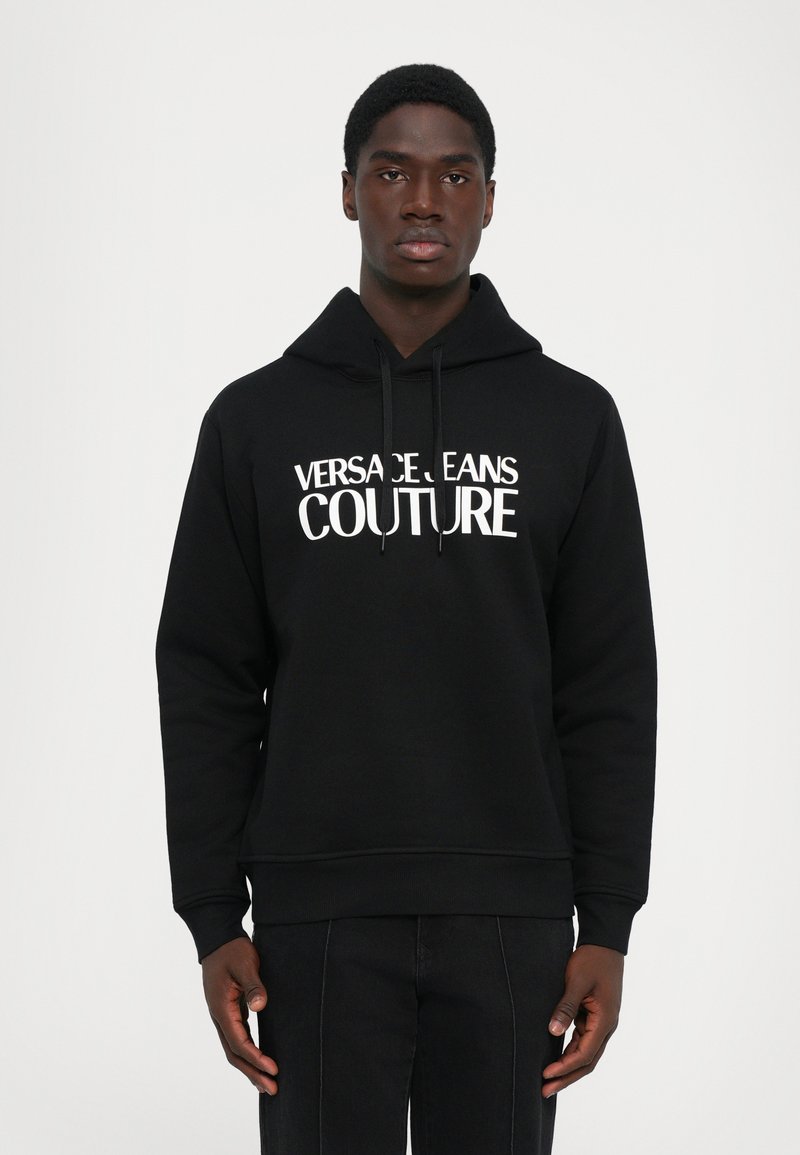 Μαύρο hoodie με λευκό κείμενο "VERSACE JEANS COUTURE" που φοριέται από έναν άνδρα που στέκεται μπροστά σε απλό λευκό φόντο, με τα χέρια χαλαρά στα πλάγια.