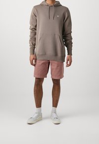 Sweat à capuche en mélange de coton taupe avec poche avant, associé à un short rose et des baskets blanches. Présente un petit logo sur la poitrine.