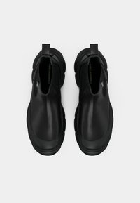 Sapatos pretos de slip-on com parte superior em couro liso, ponta arredondada, sola em borracha grossa e puxador traseiro para fácil calçar. Sem padrões visíveis.