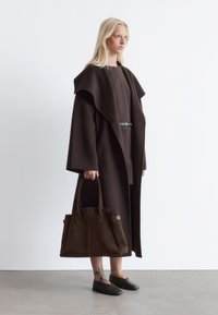 Brauner Wollmantel mit einem übergroßen Kragen, kombiniert mit einem passenden Rock und einer dunkelbraunen Wildlederhandtasche. Schwarze Schlupfschuhe und sichtbares Tattoo.