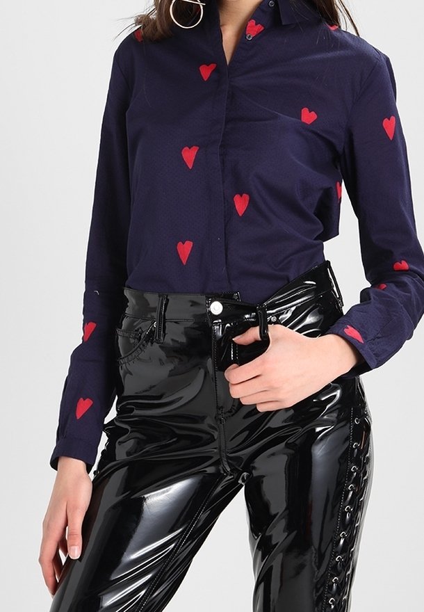 Marineblaues Hemd mit roten Herzmustern, kombiniert mit glänzenden schwarzen Vinylhosen, die einen Reißverschluss und Schnürdetails an der Seite haben.