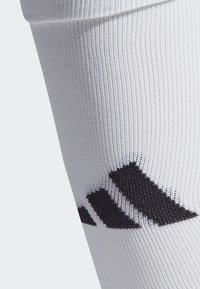 Bílá sportovní ponožka s texturovaným pleteným vzorem a černým detailem loga se třemi pruhy blízko horní části.