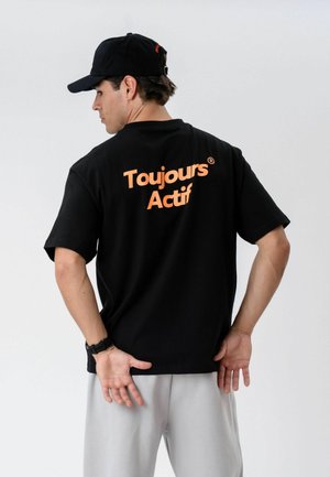 Man wearing black cap and black T-shirt with orange text "Toujours Actif" on back, standing against plain white background.