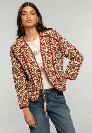 Mujer con una chaqueta de estampado floral rojo y beige sobre un top crema y jeans azules, de pie contra un fondo gris liso.