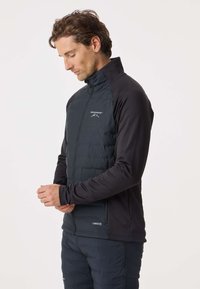 Man met bruin haar, gekleed in een zwart jack met rits en een broek, kijkt naar beneden met samengevouwen handen voor zich tegen een witte achtergrond.