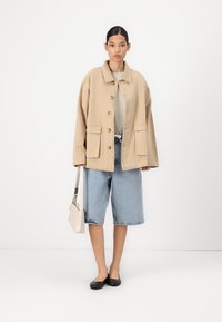 Beige oversized jacket with front buttons and pockets, paired with light blue knee-length denim shorts and black flats. Carrying a small white bag.