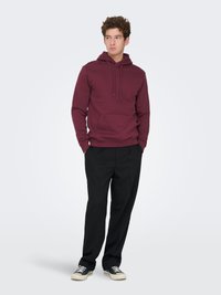 Maroon hoodie with a front pouch pocket and drawstring hood, paired with black trousers and black sneakers, featuring a textured fabric.