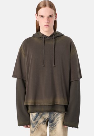 Übergroßer, zweilagiger Hoodie aus dunkelolivgrünem Baumwollmischgewebe mit Kapuze und rohen Säumen. Kombiniert mit hellen gemusterten Jeans.