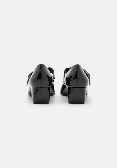 Zapatos de charol negro con un bajo tacón cuadrado, que presentan una punta afilada y una correa decorativa en el lateral. Superficie lisa y brillante.