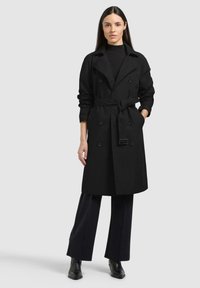 Donna con capelli lunghi e scuri, indossa un trench nero con cintura, dolcevita nero, pantaloni a gamba larga e stivaletti neri, in piedi con una mano in tasca.