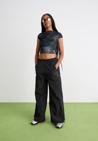 Fekete, oversize cargo nadrág oldalsó zsebekkel; sötétzöld és kék mintás, testhezálló crop top-pal párosítva. A modellen elegáns napszemüveg van.