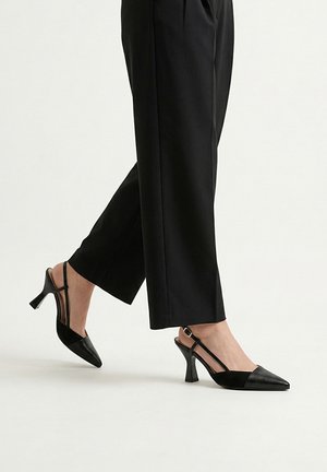 Piernas con pantalones negros de pierna ancha y tacones altos slingback negros con punteras puntiagudas sobre un fondo blanco liso.