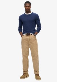 Maglione blu navy a girocollo, pantaloni beige a vestibilità rilassata e stivali marrone chiaro. Il modello sta in piedi con le mani in tasca.