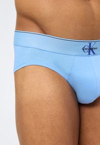 Κοντινό πλάνο ενός ατόμου που φοράει ανοιχτό μπλε εσώρουχα Calvin Klein, εμφανίζοντας τη μέση, το άνω μέρος του μηρού και τη λεπτομέρεια της ζώνης με τη μάρκα.