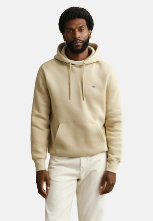 Partainen mies, joka pukeutunut beigeen huppariin, jossa on etutasku, ja valkoisiin housuihin, seisoo yksinkertaisen vaalean taustan edessä.