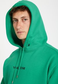 Groene katoenen hoodie met een grote capuchon, verstelbare trekkoorden en zwarte "VOLCOM" borduursels aan de voorkant. Soepele textuur, casual ontwerp.