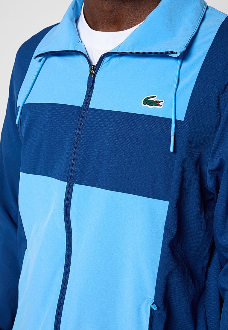 Uomo che indossa una giacca con zip blu e navy con il logo di un coccodrillo sul petto sopra una camicia bianca.