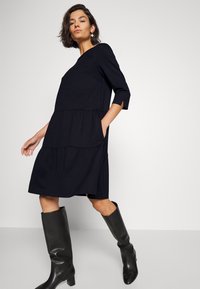 Schwarzes, gestuftes Kleid mit 3/4-Ärmeln, das seitliche Taschen und eine lockere Passform bietet. Getragen mit kniehohen schwarzen Stiefeln. Glatte Textur.