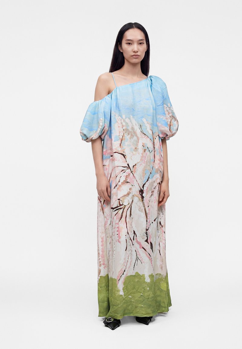 Woman wearing a long pastel floral dress with puffed sleeves, one sleeve off shoulder, standing against a plain white background.
