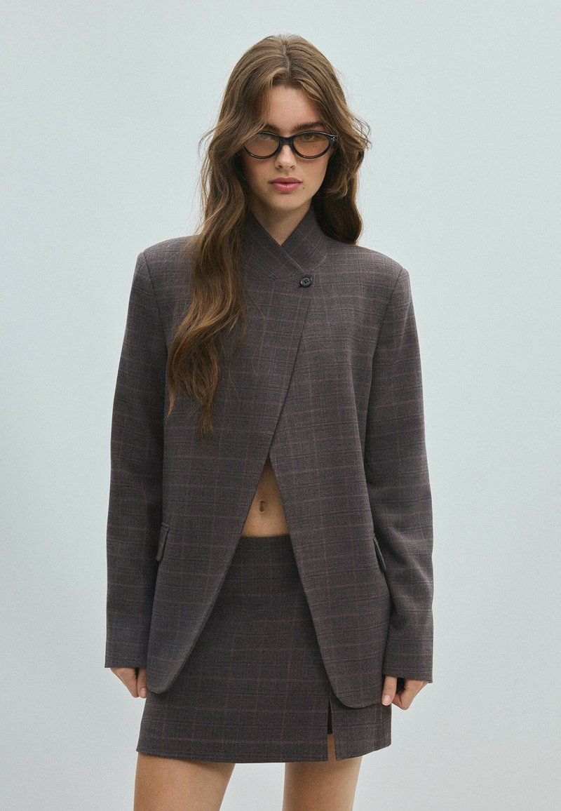 Femme aux longs cheveux bruns portant un blazer à carreaux foncés et une jupe assortie avec une fente à l'avant, ainsi que des lunettes de soleil à monture noire sur un fond uni.