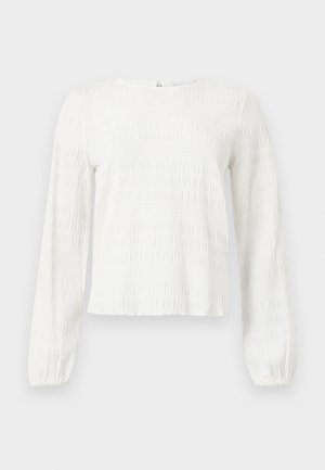 Hvit teksturert bluse med lange ermer, rund hals og elastiske mansjetter på ermene mot en ensfarget bakgrunn.