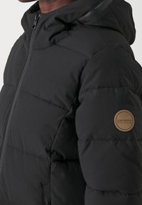 Černá péřová bunda se zipem vpředu, vysokým límcem a prošívaným designem. Vlastnosti zahrnují kruhovou hnědou nášivku na levém rukávu.