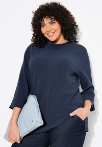 Dunkelblaues, geripptes Oberteil mit hohem Kragen und dreiviertellangen Ärmeln, kombiniert mit einer strukturierten, hellblauen Clutch. Getragen mit dunklen Denim-Hosen.