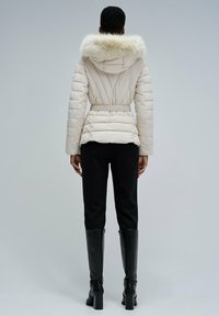 Doudoune beige clair avec capuche bordée de fourrure, taille élastique et design matelassé. Associée à un pantalon noir ajusté et des bottes à talons hauts.