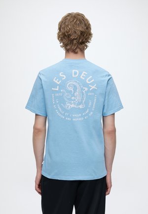 Personne aux cheveux blonds bouclés porte un t-shirt bleu clair avec un crocodile blanc et un texte en français au dos.