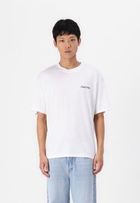 Camiseta de algodón blanca con cuello redondo, mangas cortas y un pequeño logotipo negro "Club1990" en el lado izquierdo del pecho. Llevada con jeans de color azul claro.