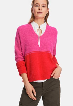 Rosa und rote V-Ausschnitt-Pullover mit lockerer Passform, gerippten Bündchen und einer weichen Textur, über einer weißen Bluse getragen.