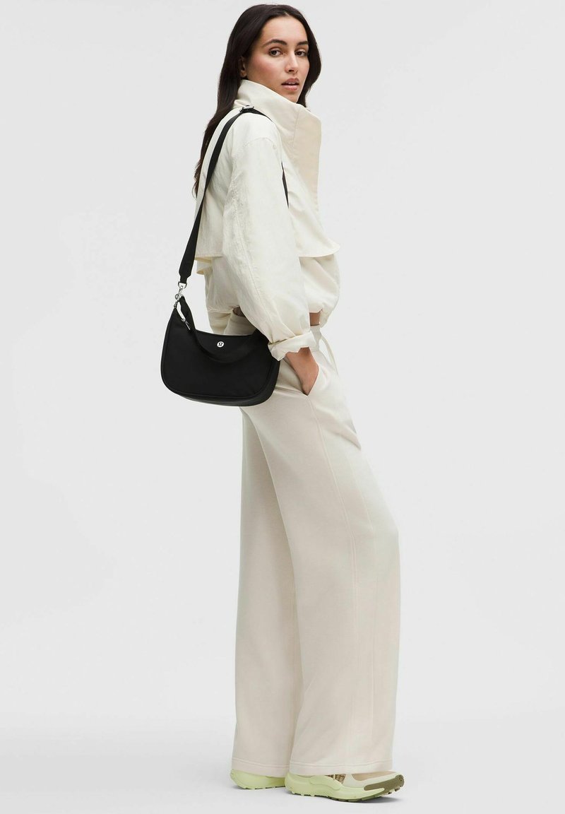 Femme en pantalon large et veste couleur écru, portant des baskets vert clair et portant un sac à bandoulière noir avec un logo argenté.