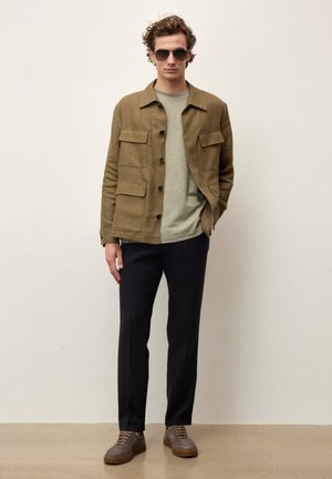 Mladi moški z sončnimi očali, rjavo jakno z žepi, svetlo sivo srajco, temnimi hlačami in rjavimi supergami stoji pred enobarvno ozadje.