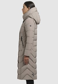 Žena v bočnom profile oblečená v dlhej taupe prešívanej vetrovke s kapucňou a vreckom so zipsom na boku, na svetlosivom pozadí.