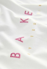 Weißes Baumwoll-T-Shirt mit pinkem Glitzer und goldfarbener Folienbeschriftung "BAKED by TED". Weicher Stoff mit glatter Oberfläche.