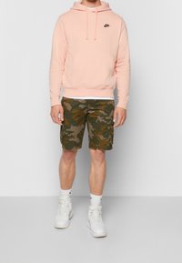 Ljust rosa Nike-hoodie med svart logotyp, camo cargoshorts i gröna nyanser och vita höga sneakers. Avslappnad sportig klädsel.