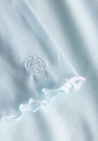 Hellblauer Stoff mit einem gerafften Rand und einem weißen, runden Sticklogo mit geometrischem Design. Glatte Textur, weiches Aussehen.