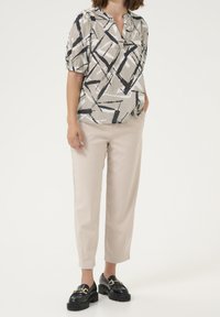 Kvinde iført en beige bluse med abstrakt mønster og korte ærmer, lyse beige cropped bukser og sorte loafers med guldbuckle detaljer.